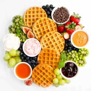 A round marble board filled with a spread of: waffles, strawberries, blueberries, kiwi, grapes, chocolate chips, peanut butter, jelly, cream cheese, and maple syrup.