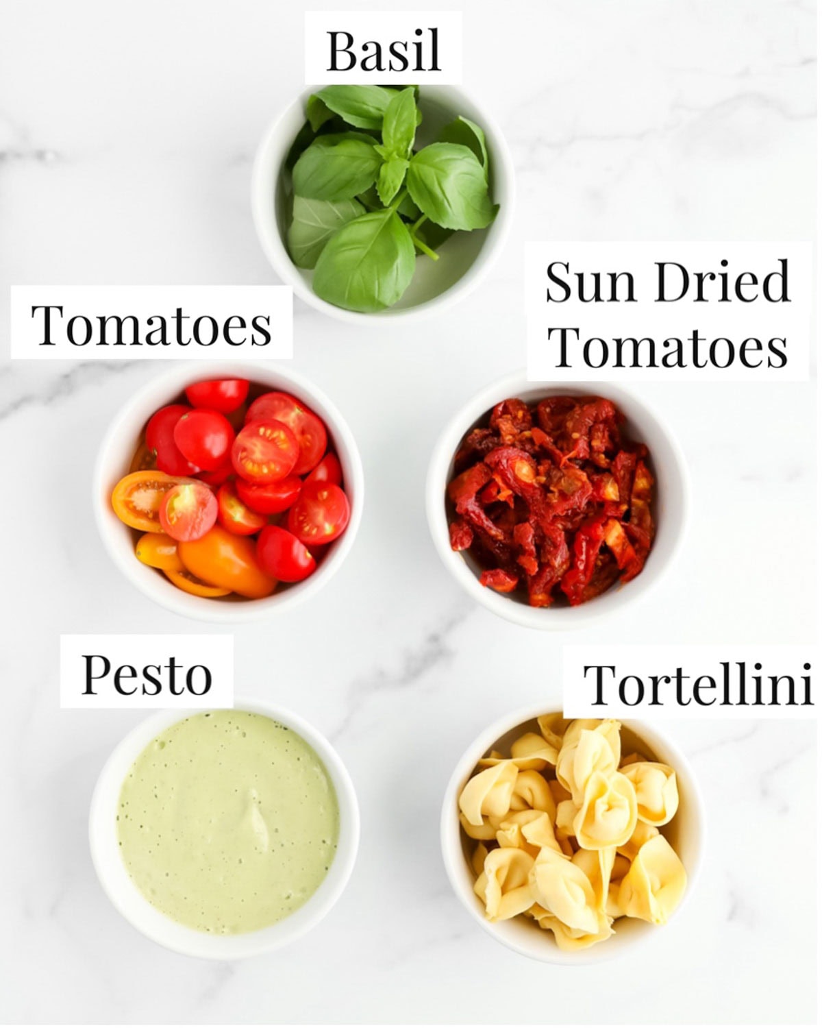 Labeled ingredients in small white bowls including: basil, tomatoes, sun dried tomatoes, pesto, and tortellini.
